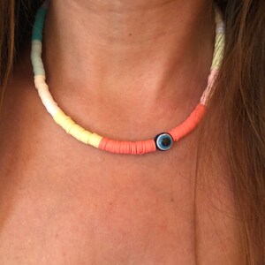 May include: A colorful beaded necklace with a blue evil eye charm. The beads are in shades of light blue, yellow, and orange.