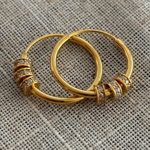 K&ouml;nnte beinhalten: Ein Paar goldene Creolen. Jeder Ohrring hat eine Reihe kleiner, funkelnder Steine, die in einem Spiraldesign am unteren Teil des Reifens angeordnet sind. Die Ohrringe werden auf einer strukturierten, neutralfarbenen Oberfl&auml;che gezeigt.