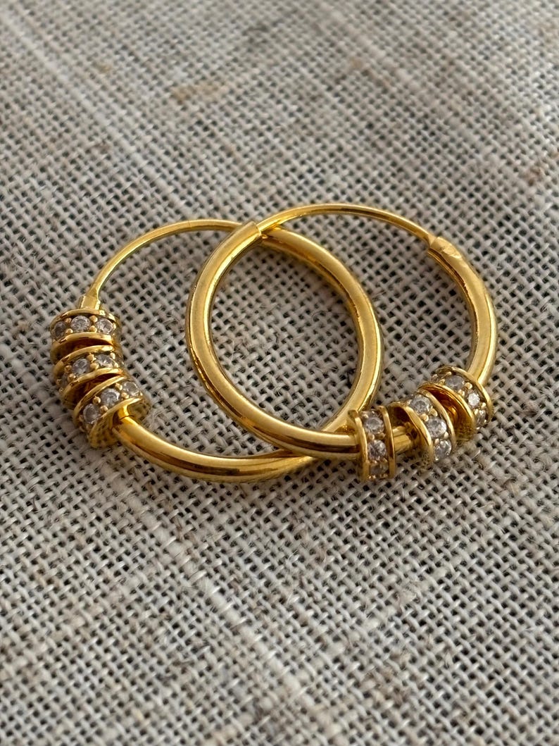 K&ouml;nnte beinhalten: Ein Paar goldene Creolen. Jeder Ohrring hat eine Reihe kleiner, funkelnder Steine, die in einem Spiraldesign am unteren Teil des Reifens angeordnet sind. Die Ohrringe werden auf einer strukturierten, neutralfarbenen Oberfl&auml;che gezeigt.