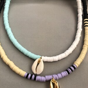 May include: A layered necklace with white, light blue, cream, and purple beads. The necklace has two shell charms, one white and one gold.