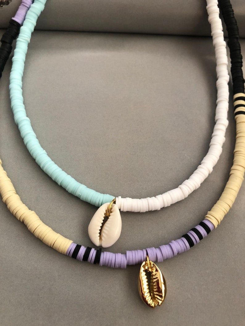 May include: A layered necklace with white, light blue, cream, and purple beads. The necklace has two shell charms, one white and one gold.