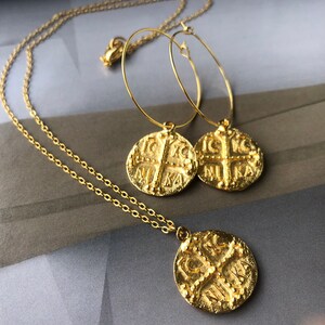 May include: Gold necklace and earrings set with a coin pendant featuring a cross and the word "NIKA" in Greek. The necklace has a delicate chain and the earrings are hoop style.