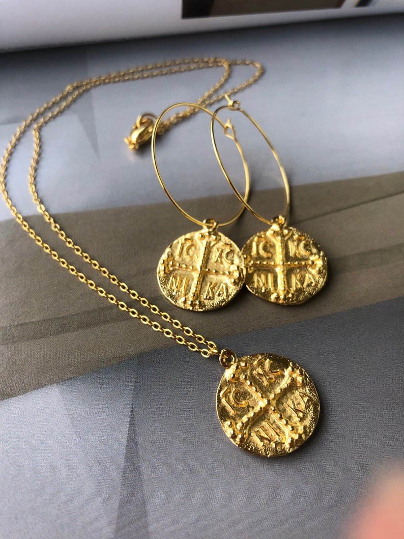 May include: Gold necklace and earrings set with a coin pendant featuring a cross and the word "NIKA" in Greek. The necklace has a delicate chain and the earrings are hoop style.