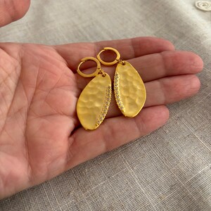 May include: Gold-colored, hammered-texture earrings with a teardrop shape. Each earring features a row of small, clear stones along one side. The earrings are held by a person's hand, showcasing their design and craftsmanship.