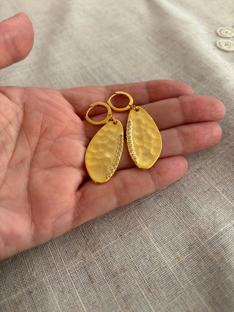 May include: Gold-colored, hammered-texture earrings with a teardrop shape. Each earring features a row of small, clear stones along one side. The earrings are held by a person's hand, showcasing their design and craftsmanship.