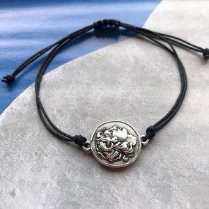 May include: A black cord bracelet with a silver circular charm featuring a detailed profile of a face.