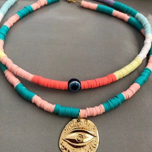 May include: A layered necklace with colorful beads and a gold pendant featuring an evil eye design. The necklace is made of pink, blue, white, and yellow beads.
