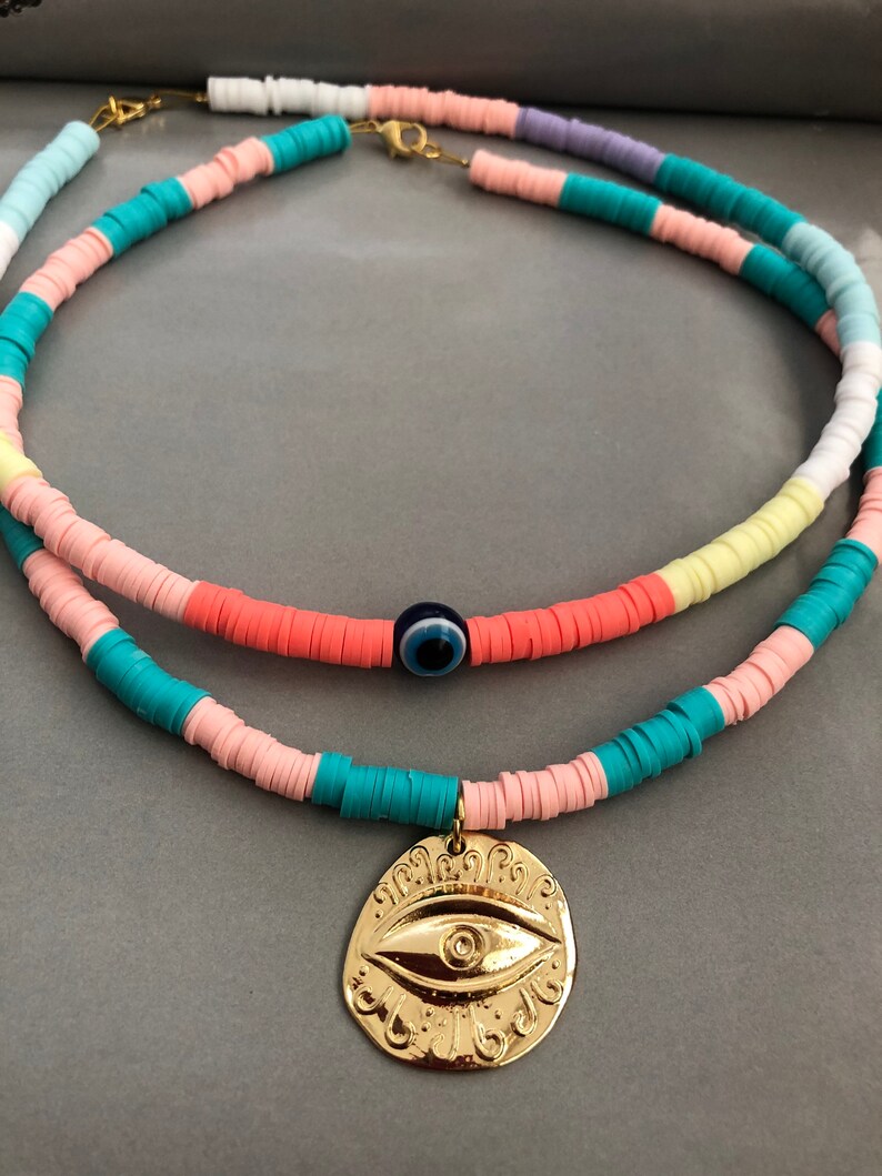 May include: A layered necklace with colorful beads and a gold pendant featuring an evil eye design. The necklace is made of pink, blue, white, and yellow beads.