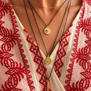 May include: A layered necklace set featuring a thin gold chain and three black cords. The cords hold gold-toned pendants: a circular charm with a small dark eye motif, a teardrop-shaped charm with a star design, and a long, slender vertical bar.