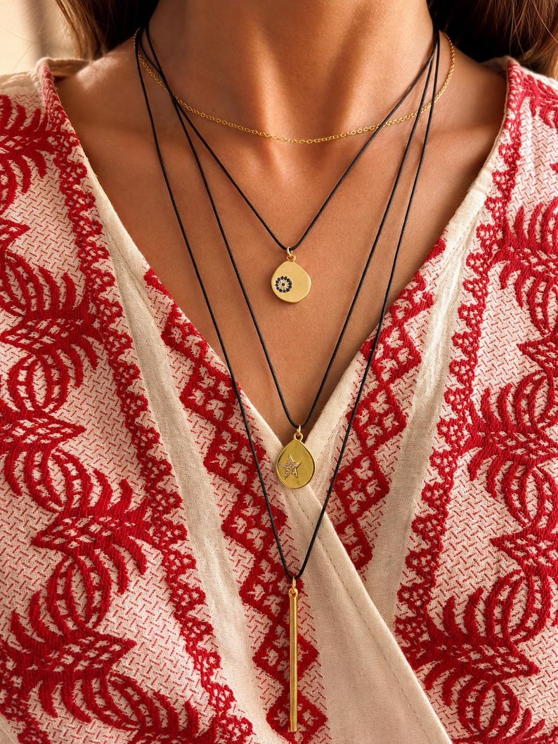 May include: A layered necklace set featuring a thin gold chain and three black cords. The cords hold gold-toned pendants: a circular charm with a small dark eye motif, a teardrop-shaped charm with a star design, and a long, slender vertical bar.
