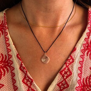 May include: A layered necklace set consisting of a thin gold-tone metal chain and a black cord necklace. The black cord features a small, irregular, light-colored pendant with a textured, heart-shaped detail in the center of the charm.