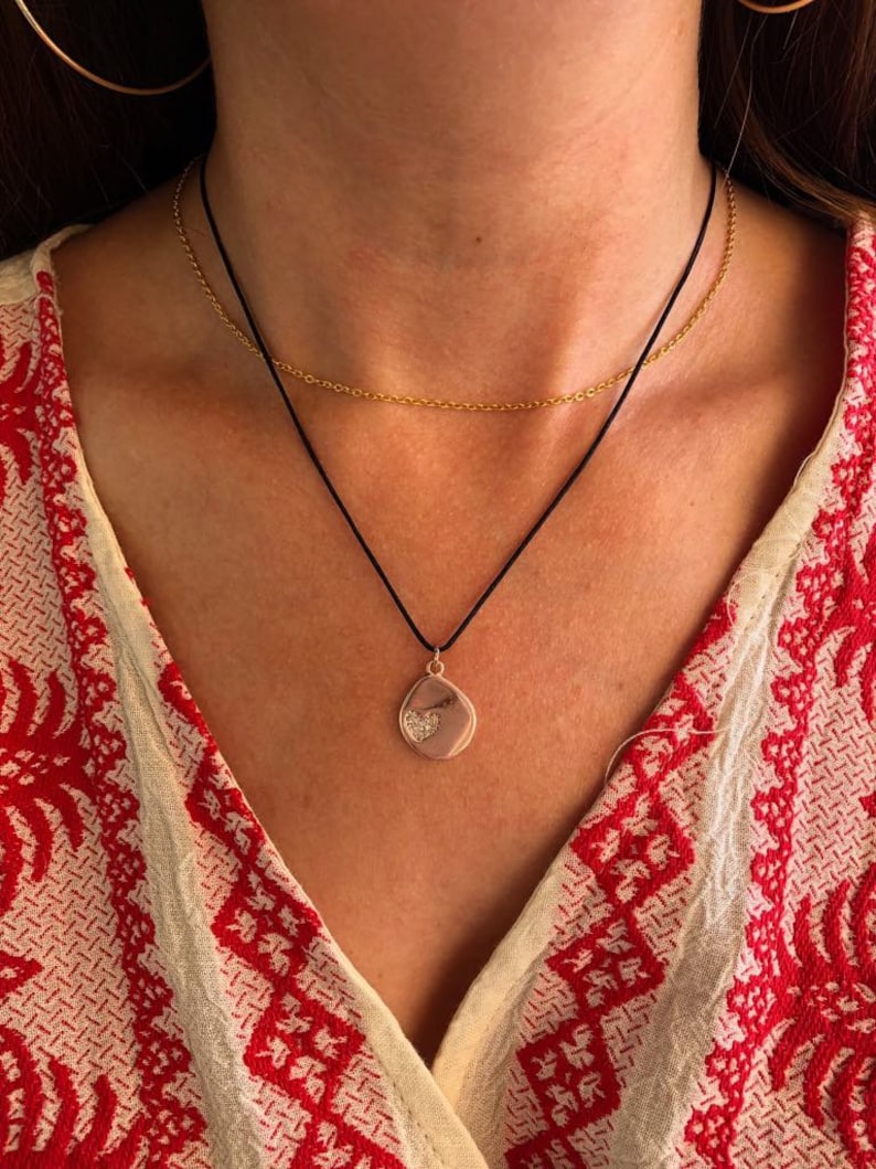 May include: A layered necklace set consisting of a thin gold-tone metal chain and a black cord necklace. The black cord features a small, irregular, light-colored pendant with a textured, heart-shaped detail in the center of the charm.