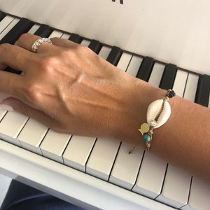 May include: A gold bracelet with a white seashell charm, turquoise bead, and gold fish charm. The bracelet is on a person's wrist, resting on a white piano keyboard.