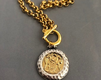 Puede incluir: Un collar de cadena dorada con un colgante de plata y oro con un diseño en relieve de dos figuras. El colgante tiene una textura martillada.