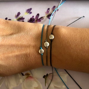 Puede incluir: Tres pulseras de la amistad con dijes plateados en forma de sol con un ojo en el centro. Las pulseras están atadas en la muñeca de una persona. Las pulseras son azules, grises y negras.