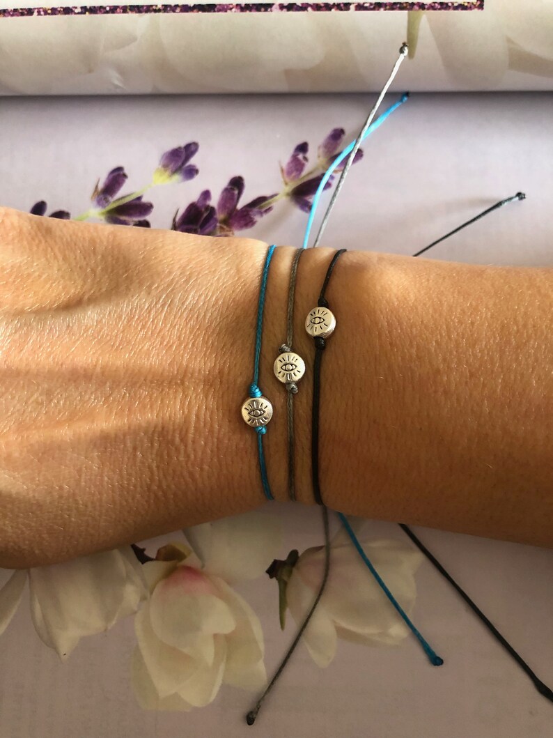 May include: Three friendship bracelets with silver charms in the shape of a sun with an eye in the center. The bracelets are tied on a person's wrist. The bracelets are blue, gray, and black.