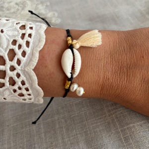 Peut inclure: Un bracelet en cordon noir avec un coquillage blanc, des perles dorées et une petite gland crème. Le bracelet comprend également deux petites perles. Ce bijou fait main est un accessoire élégant.