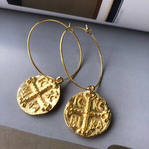 May include: Gold hoop earrings with a round coin charm featuring a cross and the inscription "IC XC NIKA".