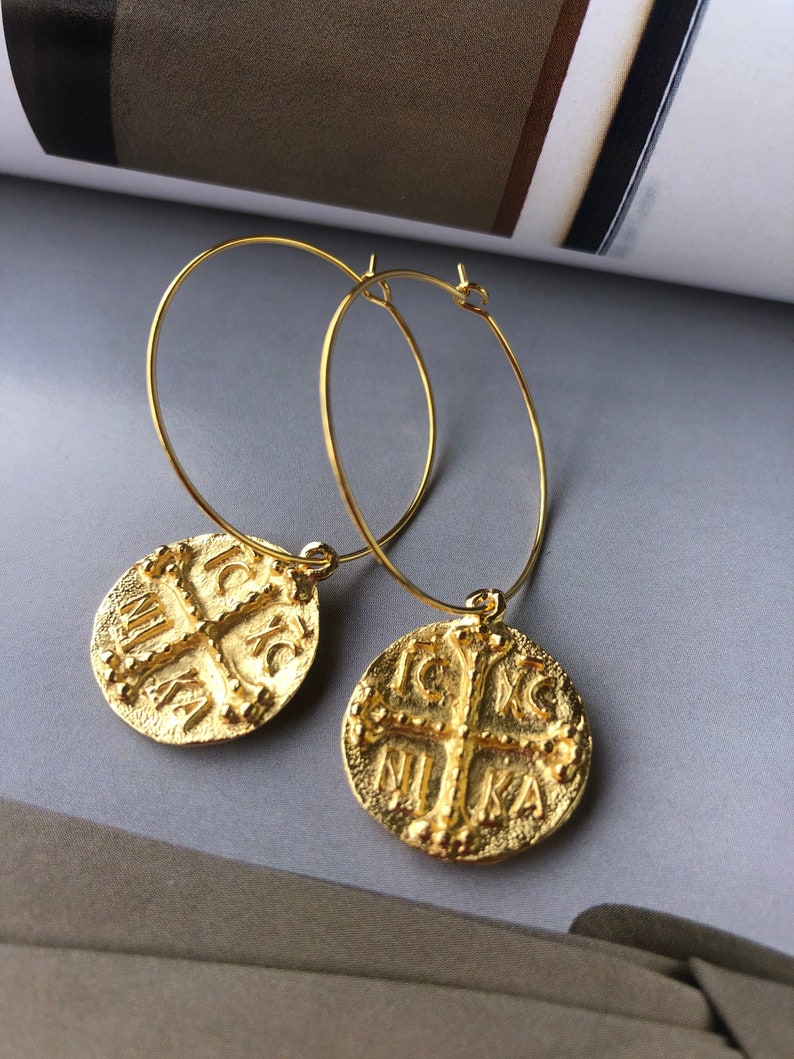 May include: Gold hoop earrings with a round coin charm featuring a cross and the inscription "IC XC NIKA".