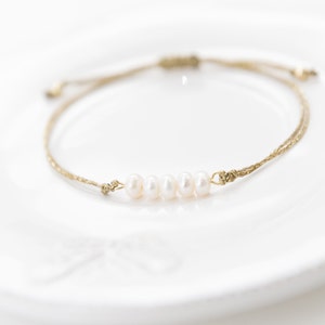 May include: A simple bracelet made with a brown cord and five white pearls. The bracelet is adjustable and can be worn on the wrist or ankle.