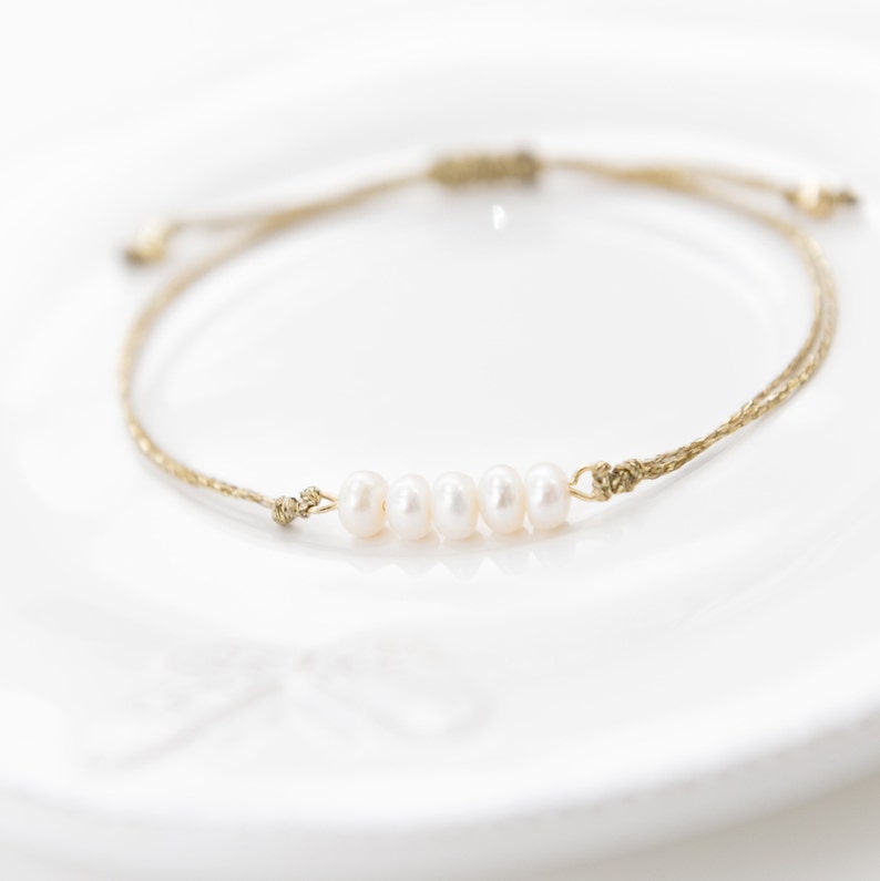 May include: A simple bracelet made with a brown cord and five white pearls. The bracelet is adjustable and can be worn on the wrist or ankle.
