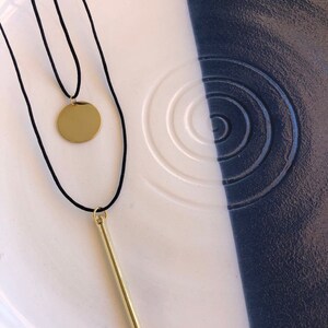 May include: A layered necklace featuring two black cords. One cord holds a small, polished gold-tone circular pendant, while the other cord features a long, slender, polished gold-tone bar pendant. Both pendants hang vertically from the black cords.