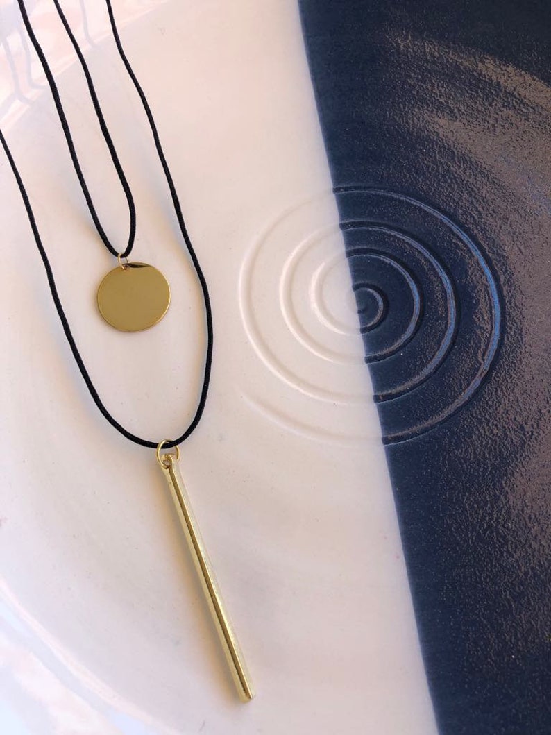 May include: A layered necklace featuring two black cords. One cord holds a small, polished gold-tone circular pendant, while the other cord features a long, slender, polished gold-tone bar pendant. Both pendants hang vertically from the black cords.
