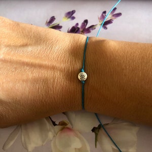 May include: A turquoise blue string bracelet with a silver charm featuring a sun design.