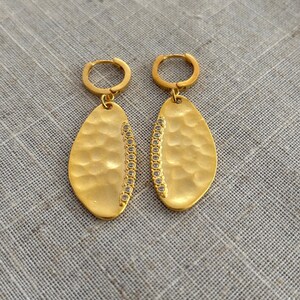 May include: A pair of gold-colored earrings with a hammered texture and a row of small, clear stones along one side. The earrings have a teardrop shape and are suspended from small hoop closures.
