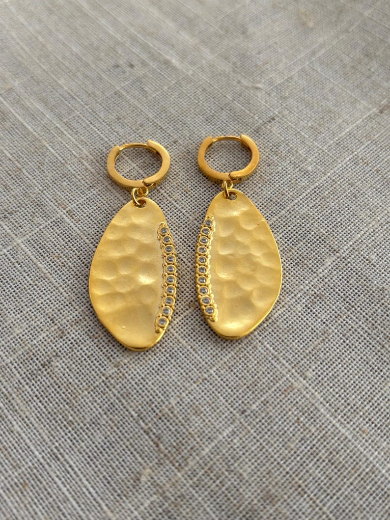 May include: A pair of gold-colored earrings with a hammered texture and a row of small, clear stones along one side. The earrings have a teardrop shape and are suspended from small hoop closures.
