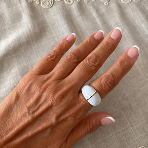 Könnte beinhalten: Ein breiter, glatter weißer Ring mit einem dünnen, horizontalen goldfarbenen Banddetail in der Mitte. Der Ring hat eine abgerundete, gewölbte Form und eine glänzende Oberfläche, die den Finger mit einem klaren, soliden Aussehen umschließt.