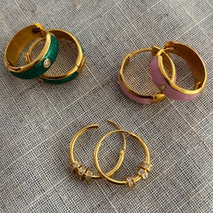 K&ouml;nnte beinhalten: Drei Paar goldfarbene Creolen. Ein Paar hat gr&uuml;ne Emaille und einen kleinen Diamanten. Ein weiteres Paar hat rosa Emaille und einen kleinen Diamanten. Das dritte Paar hat kleine klare Steine.