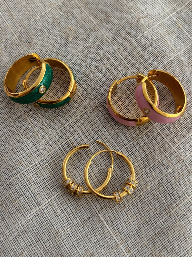 K&ouml;nnte beinhalten: Drei Paar goldfarbene Creolen. Ein Paar hat gr&uuml;ne Emaille und einen kleinen Diamanten. Ein weiteres Paar hat rosa Emaille und einen kleinen Diamanten. Das dritte Paar hat kleine klare Steine.