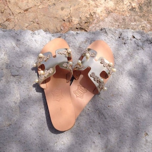 May include: Gold leather sandals with pearl embellishments and delicate floral details. The sandals have a simple, slip-on design and a comfortable flat sole.