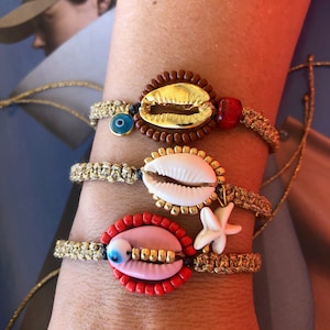 May include: Three bracelets with gold accents and various charms. The top bracelet has a brown shell charm with gold accents and a blue evil eye charm. The middle bracelet has a white shell charm with gold accents and a gold starfish charm. The bottom bracelet has a pink shell charm with gold accents and a blue evil eye charm.