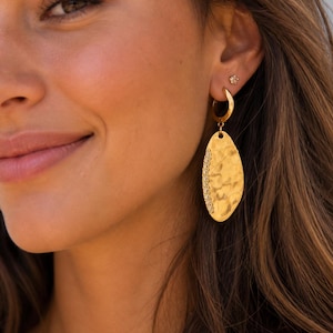 May include: Close-up of a woman wearing gold earrings. The earrings have a hammered texture and an oval shape, with a row of small stones along the edge. A small gold stud earring is also visible.