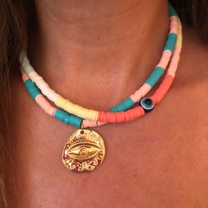 May include: A colorful layered beaded necklace with a large gold evil eye pendant. The necklace is made of small, round beads in shades of pink, blue, yellow, and white. The pendant is a circular disc with a detailed evil eye design.