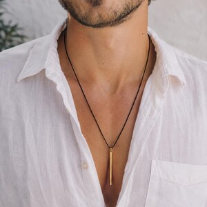 May include: A gold-toned cylindrical pendant suspended from a thin black cord necklace. The pendant is a long, slender, straight metal bar with a smooth finish, hanging vertically against a white button-down shirt with a visible chest pocket.