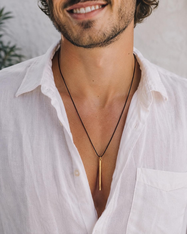 May include: A gold-toned cylindrical pendant suspended from a thin black cord necklace. The pendant is a long, slender, straight metal bar with a smooth finish, hanging vertically against a white button-down shirt with a visible chest pocket.