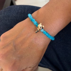Puede incluir: Una pulsera elástica compuesta por pequeñas cuentas redondas y facetadas de color azul turquesa. La pulsera presenta un dije central dorado con forma de cabeza de gato, con un dije más pequeño y colgante de espina de pescado dorado debajo.