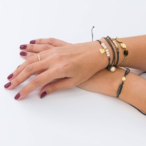Puede incluir: Una muñeca de mujer adornada con varios brazaletes. Los brazaletes están hechos de cordón negro y presentan detalles dorados, perlas y un dije en forma de corazón.