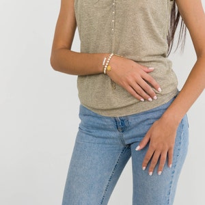 May include: A woman wearing a light brown sleeveless top and blue jeans. She is wearing a gold bracelet with a pearl charm.