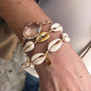 May include: A layered bracelet with a large clear quartz crystal, gold shell charms, and white cowrie shells on a brown cord.