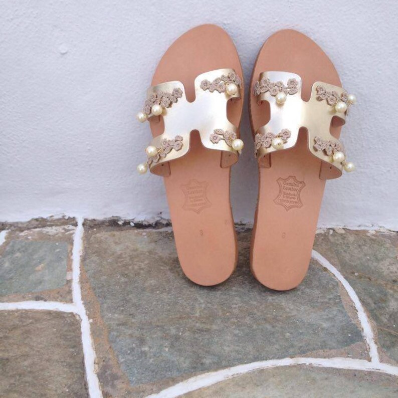 May include: Gold leather sandals with a delicate lace design and pearl embellishments. The sandals have a simple, flat sole and are made in Greece.