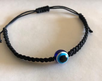 May include: A black string bracelet with a blue evil eye charm. The charm has a white circle around the blue eye and a black pupil.