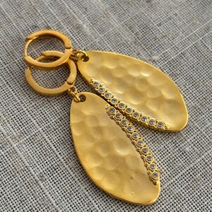 May include: A pair of gold-colored oval earrings with a hammered texture. Each earring features a line of small, clear gemstones along one side. The earrings have a hoop closure.