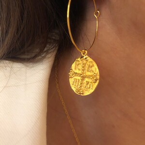 May include: Gold hoop earrings with a round coin charm featuring a cross and the letters 'IXKA'.