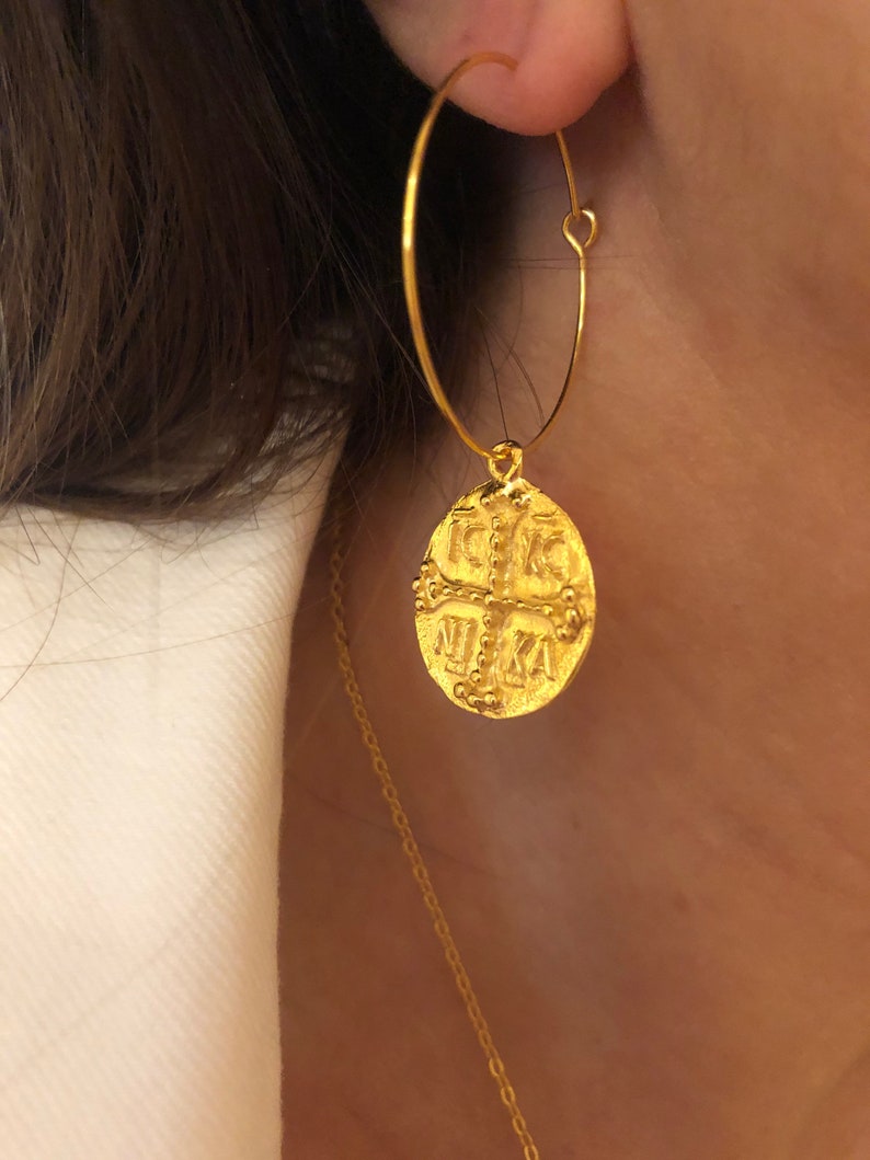 May include: Gold hoop earrings with a round coin charm featuring a cross and the letters 'IXKA'.