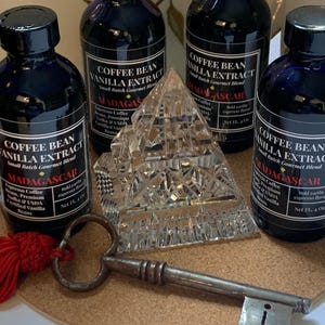 May include: Four small glass bottles of coffee bean vanilla extract with black labels and gold lettering. The bottles are arranged in a pyramid shape on a cork surface. The labels read "Coffee Bean Vanilla Extract, Small Batch Gourmet Blend, Madagascar". A crystal pyramid and an antique key are also on the surface.