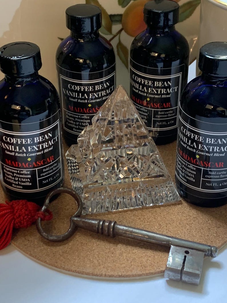 May include: Four small glass bottles of coffee bean vanilla extract with black labels and gold lettering. The bottles are arranged in a pyramid shape on a cork surface. The labels read "Coffee Bean Vanilla Extract, Small Batch Gourmet Blend, Madagascar". A crystal pyramid and an antique key are also on the surface.